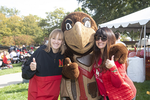 two women standing with Testudo mascot in the center. All have their thumbs up.